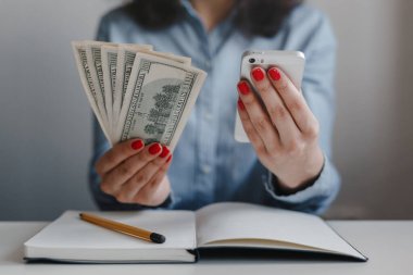 Yüz dolarlık banknotlar tutan kırmızı tırnaklı kadınların elleri ve masada oturan mavi gömlekli bir cep telefonu. İş, serbest meslek, uzak iş, online iş kavramı