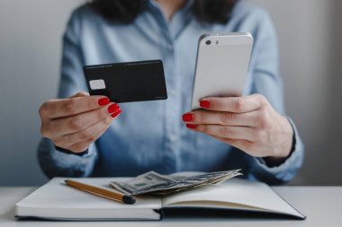 Kırmızı tırnaklı, elinde kredi kartı ve telefon olan mavi gömlekli bir kadının masanda oturup ödemesini yaptığı fotoğraf. İş, serbest meslek, uzak iş, online iş kavramı