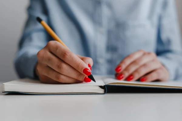 Closeup of woman\'s hands with red nails making notes in a notebook at the desk. Wearing blue shirt at work place. Business, freelance, self-employment. Distance job and online work concept