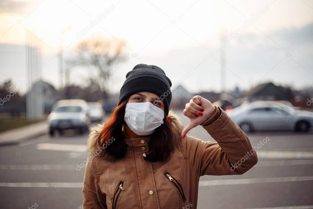 Sick unhappy girl, young woman wearing surgery medical mask feeling bad, ill, being hit by the coronavirus, Covid2019 holds hand shows dislike thumbs down fingers gesture. Worldwide pandemic concept