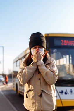 Genç bir kadın otobüs durağında hapşırıyor ve yüzünü ellerle kapatıyor. Toplu taşıma istasyonunda bir otobüsün yanında duran beyaz bir tıbbi maske takıyor. Coronavirus hastalığı konsepti. Pandemia, epidemi