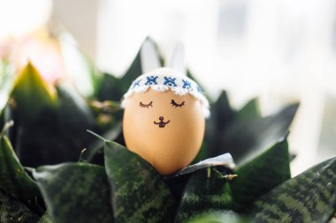 Kulak yumurtası olan küçük bir Paskalya tavşanı. Üzerinde yeşil çiçek çalılarının arasında çizilmiş bir yüz var. Paskalya tatili süslemeleri ve hazırlık konsepti. Kutsal Din Günü