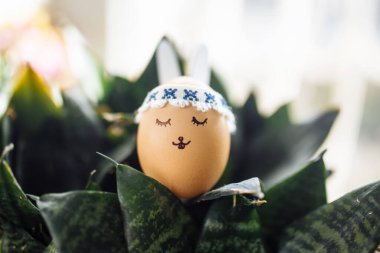 Kulak yumurtası olan küçük bir Paskalya tavşanı. Üzerinde yeşil çiçek çalılarının arasında çizilmiş bir yüz var. Paskalya tatili süslemeleri ve hazırlık konsepti. Kutsal Din Günü