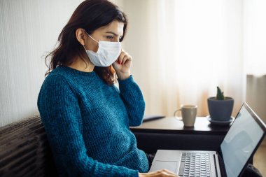 İş kadını evde kalır ve koronavirüs epidemisi karantinasında çalışır. Kadın işçi, tıbbi maskesine dokunuyor ve dizüstü bilgisayarında daktilo yazıyor. Covid-19 pandemisi yayılma önleme kavramı