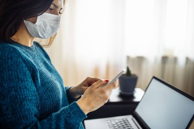 İş kadını evde kalır ve koronavirüs epidemisi karantinasında çalışır. Tıbbi maske takan ve cep telefonunu elinde tutan bir kadın işçi. Covid-19 pandemisi yayılma önleme kavramı