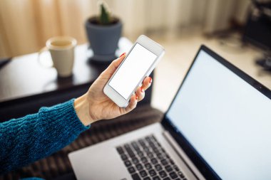 Genç bir kadının cep telefonuna yakın çekim. Evde kalıyor ve işleri uzaktan yönetiyor. Serbest olarak. Laptop, kupa ve çiçek arka planda.
