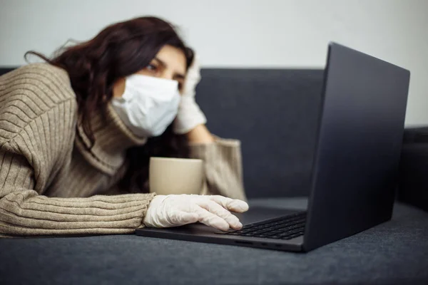 Young woman working from home during quarantine due to coronavirus pandemia. Beautiful girl stays home wearing medical mask and gloves and typing on a laptop. Covid-19 epidemia worldwide concept