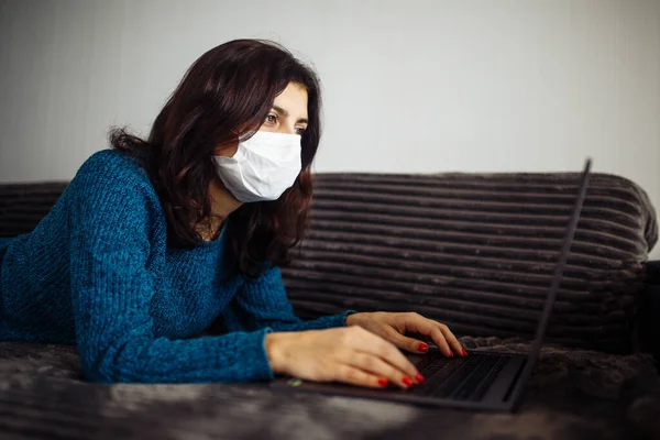 Young businesswoman working from home during quarantine due to coronavirus pandemia. Beautiful girl stays home wearing medical mask and typing on a laptop. Covid-19 epidemia worldwide concept