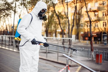 Kişisel koruyucu giysi, eldiven ve gaz maskesi takmış bir adam. Sokakları uyarı bandıyla temizliyor. Sırt çantası dolusu sprey dezenfektan ile korunuyor.