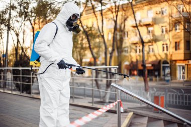 Kişisel koruyucu giysi, eldiven ve gaz maskesi takmış bir adam. Sokakları uyarı bandıyla temizliyor. Sırt çantası dolusu sprey dezenfektan ile korunuyor.