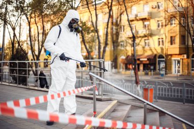 Kişisel koruyucu giysi, eldiven ve gaz maskesi takmış bir adam. Sokakları uyarı bandıyla temizliyor. Sırt çantası dolusu sprey dezenfektan ile korunuyor.