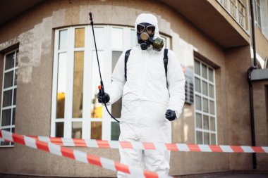 Virüs koruyucu kıyafet giyen hijyenik istasyon çalışanı elinde dezenfeksiyon spreyi ve sırt çantasıyla kırmızı ve beyaz bandın arkasında duruyor. Coronavirus covid-19 önleme