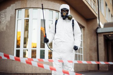 Virüs koruyucu kıyafet giyen hijyenik istasyon çalışanı elinde dezenfeksiyon spreyi ve sırt çantasıyla kırmızı ve beyaz bandın arkasında duruyor. Coronavirus covid-19 önleme