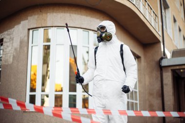 Virüs koruyucu kıyafet giyen hijyenik istasyon çalışanı elinde dezenfeksiyon spreyi ve sırt çantasıyla kırmızı ve beyaz bandın arkasında duruyor. Coronavirus covid-19 önleme