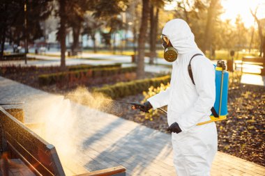 Özel korumalı dezenfeksiyon kıyafeti giyen bir adam şehirde yayılan koronavirüsün ortasında boş bir parktaki bankın üzerine sterilizatör sıkar. Güneşli bir arka plan. Dünya çapında Covid-19 'u durdurun.