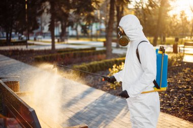Özel korumalı dezenfeksiyon kıyafeti giyen bir adam şehirde yayılan koronavirüsün ortasında boş bir parktaki bankın üzerine sterilizatör sıkar. Güneşli bir arka plan. Dünya çapında Covid-19 'u durdurun.
