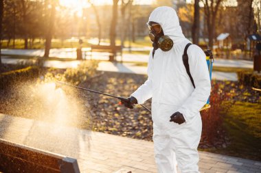 A man wearing special protective disinfection suit sprays sterilizer on a bench in the empty park to amid coronavirus spread in the city. Sunny background. Stop Covid-19 worldwide clipart