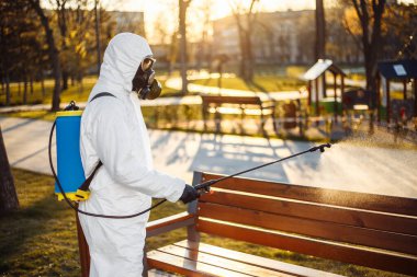 Özel korumalı dezenfeksiyon kıyafeti giyen bir adam şehirde yayılan koronavirüsün ortasında boş bir parktaki bankın üzerine sterilizatör sıkar. Güneşli bir arka plan. Dünya çapında Covid-19 'u durdurun.