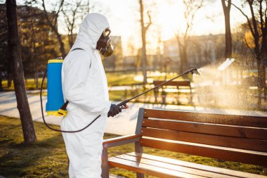 Özel korumalı dezenfeksiyon kıyafeti giyen bir adam şehirde yayılan koronavirüsün ortasında boş bir parktaki bankın üzerine sterilizatör sıkar. Güneşli bir arka plan. Dünya çapında Covid-19 'u durdurun.
