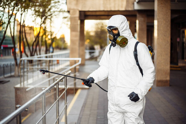 Man sprays disinfector onto the railing wearing coronavirus protective suit and equipment. Cleaning and sterilizing the not crowded city streets. Covid-19 nCov2019 spread prevention