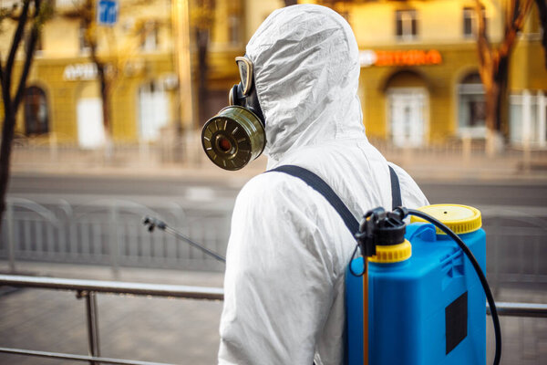 Man sprays disinfector onto the railing wearing coronavirus protective suit and equipment. Cleaning and sterilizing the not crowded city streets. Covid-19 nCov2019 spread prevention