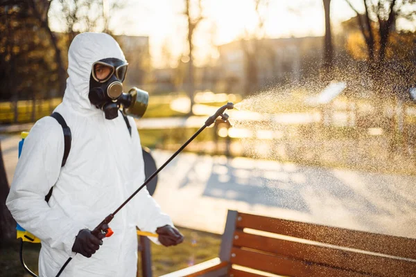 A man wearing special protective disinfection suit sprays sterilizer on a bench in the empty park to amid coronavirus spread in the city. Sunny background. Stop Covid-19 worldwide