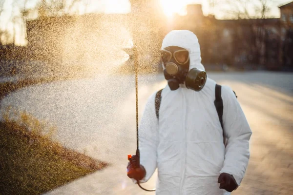 A man in a protective disinfection suit sprays sterilizer on the park square to prevent spread of coronavirus covid-19. Professional saniraty worker cleans city streets from the virus. Spot nCov2019