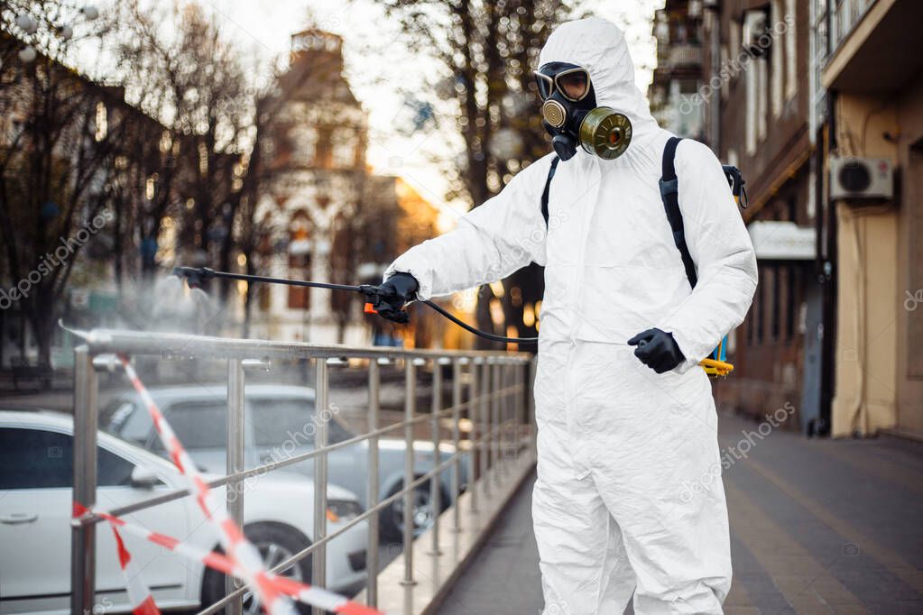 A man in protective equipment disinfects with a sprayer in the city. Surface treatment due to coronavirus covid-19 disease. A man in a white suit disinfects the street and rails. Virus pandemic