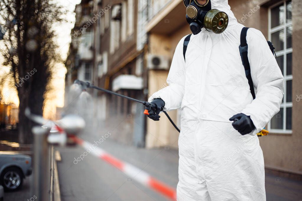 A man in protective equipment disinfects with a sprayer in the city. Surface treatment due to coronavirus covid-19 disease. A man in a white suit disinfects the street and rails. Virus pandemic