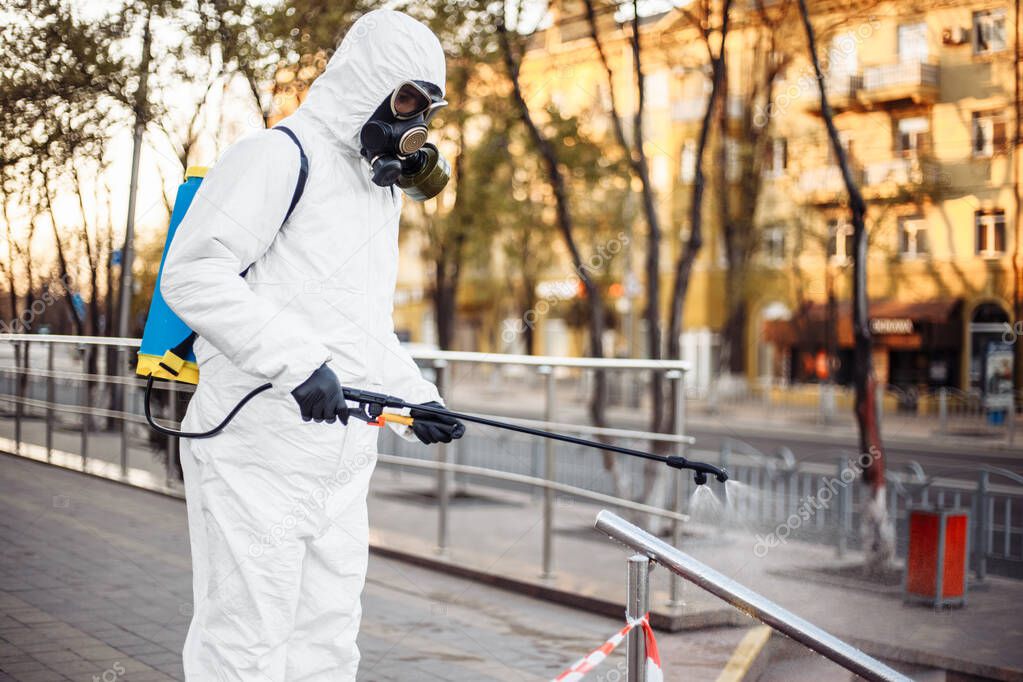 Man wearing personal protective equipment suit, gloves and gas mask cleaning the streets protected with warning tape with a backpack of spray disinfectant water to remove covid-19 coronavirus