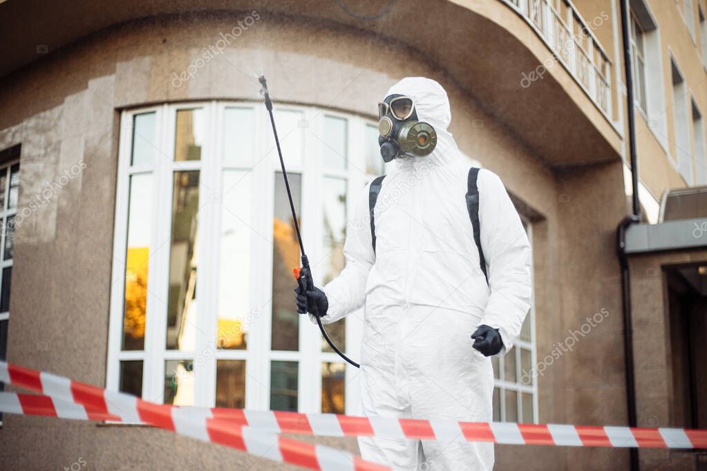 The worker of sanitary station wearing virus protective suit stands behind warning red and white tape with a disinfetion spray equipment in the hand and backpack. Coronavirus covid-19 prevention