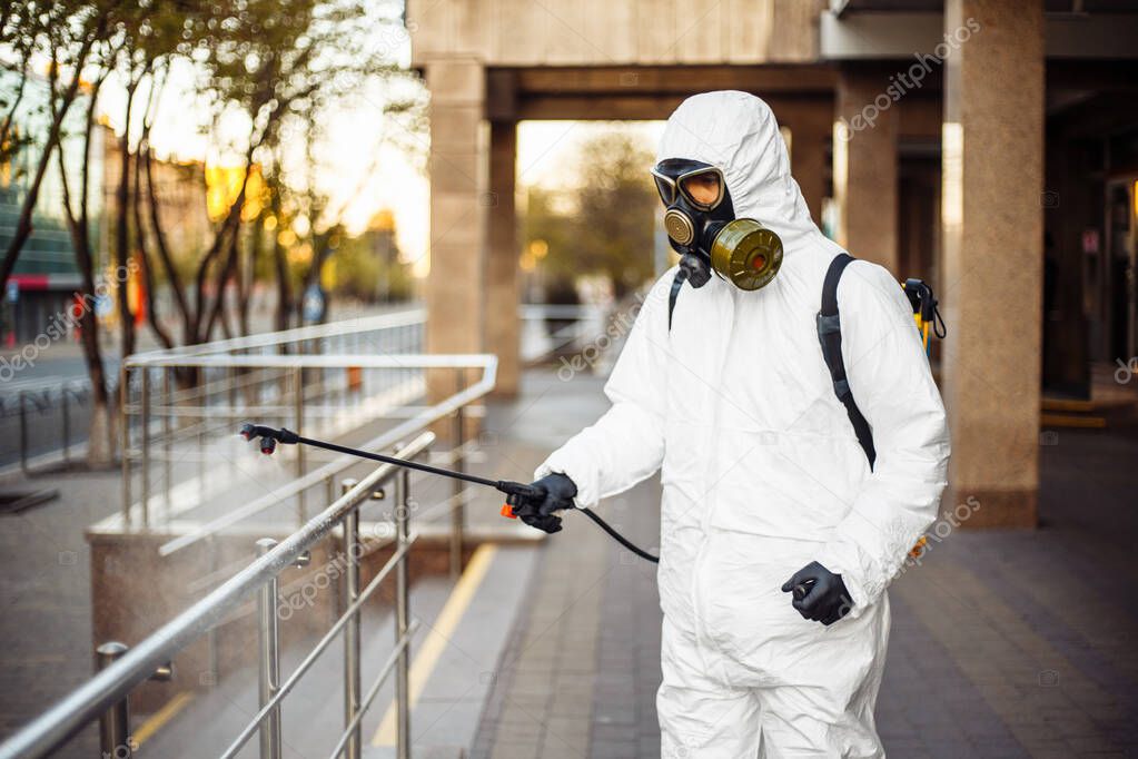 Man sprays disinfector onto the railing wearing coronavirus protective suit and equipment. Cleaning and sterilizing the not crowded city streets. Covid-19 nCov2019 spread prevention