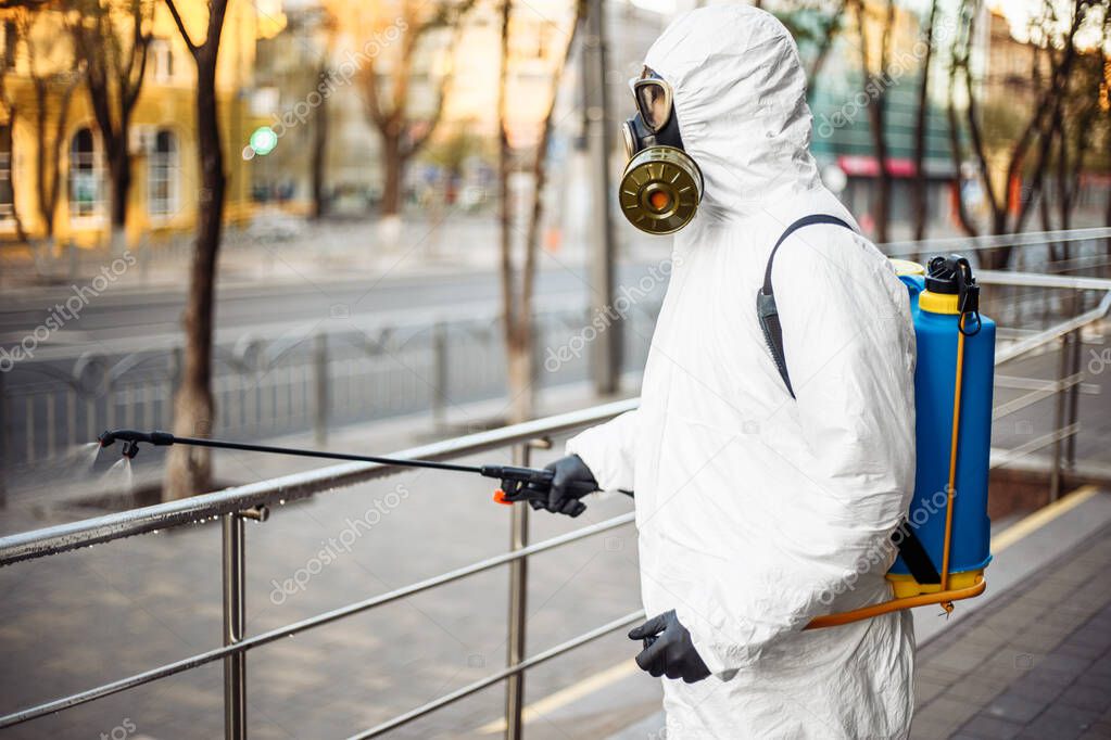 Man sprays disinfector onto the railing wearing coronavirus protective suit and equipment. Cleaning and sterilizing the not crowded city streets. Covid-19 nCov2019 spread prevention