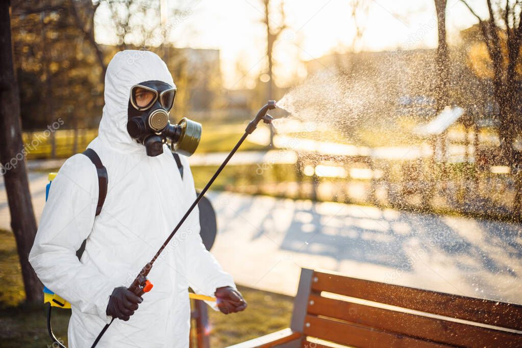 A man wearing special protective disinfection suit sprays sterilizer on a bench in the empty park to amid coronavirus spread in the city. Sunny background. Stop Covid-19 worldwide
