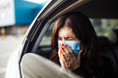 Arka koltukta tıbbi steril maske takan bir kadın ve elleriyle pencereden dışarı hapşırıyor. Coronavirus karantinası sırasında trafik sıkışıklığında kalmış bir kız yolcu. Sağlık hizmetleri kavramı
