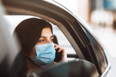 Taksinin arka koltuğunda tıbbi steril maske takan bir kadın camdan dışarı bakıp telefonla konuşuyor. Coronavirus karantinası sırasında trafik sıkışıklığında bekleyen kız yolcu. Sağlık hizmetleri kavramı