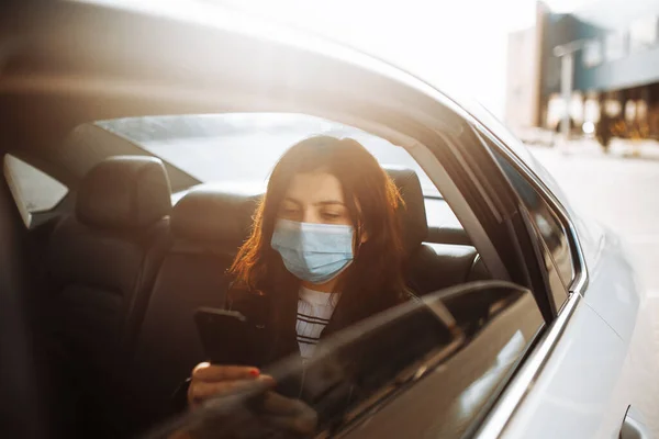 Taksi arabasında steril bir maske takan bir kadın arka koltukta camdan dışarı bakarak cep telefonunu kontrol ediyor. Coronavirus karantinası sırasında trafik sıkışıklığında bekleyen kız yolcu. Sağlık hizmetleri kavramı