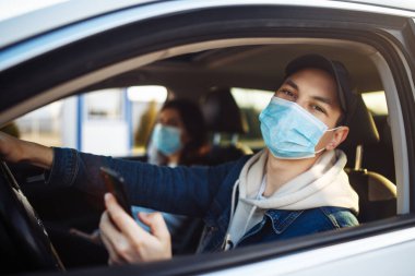Tıbbi maske takan bir adam araba sürerken cep telefonunu elinde tutar. Çocuk sürücü trafik sıkışıklığında Coronavirus Dünyası haberlerini kontrol ediyor. Karantina sırasında izolasyon ve sağlık hizmetleri