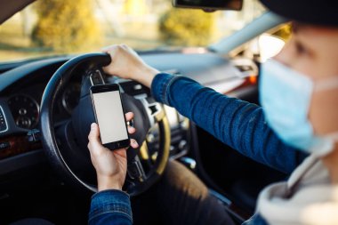 Tıbbi maske takan bir adam araba sürerken cep telefonunu elinde tutar. Çocuk sürücü trafik sıkışıklığında Coronavirus Dünyası haberlerini kontrol ediyor. Karantina sırasında izolasyon ve sağlık hizmetleri