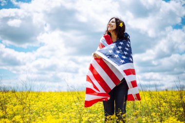 Sarı çiçekli güzel bir tarlada Amerikan bayrağına sarılmış vatansever genç bir kadın. ABD 'nin bağımsızlık günü 4 Temmuz kutlamaları. Yaz tatili kavramı