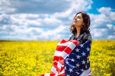 Sarı çiçekli güzel bir tarlada Amerikan bayrağına sarılmış vatansever genç bir kadın. ABD 'nin bağımsızlık günü 4 Temmuz kutlamaları. Yaz tatili kavramı