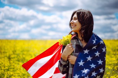 Güzel bir tarlada elinde bir buket sarı çiçekle Amerikan bayrağına sarılmış vatansever genç bir kadın. ABD 'nin bağımsızlık günü 4 Temmuz kutlamaları. Yaz tatili kavramı