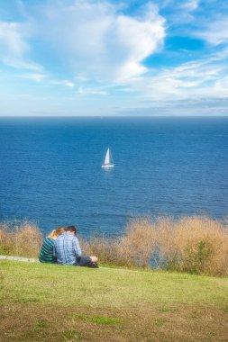 Genç çift çimenlerde oturmuş okyanus manzarasının tadını çıkarıyor.