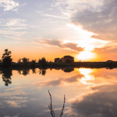 Ağaçların yansımasıyla küçük sessiz göl ve gündoğumu gökyüzü.