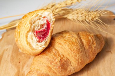 Isırılmış kruvasan ve çilek reçeli. Beyaz arka plandaki tahtanın üzerinde kuru buğday var.