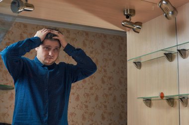 Kafkasyalı genç bir adam aynanın yanında dikiliyor. Kafasını tutuyor. Ev aletinin mavi gömleğiyle aynaya bakıyor.