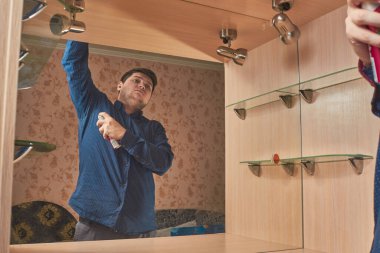 Genç beyaz bir adam, evinde mavi gömlekli bir deodorant ile aynanın yanında duruyor.