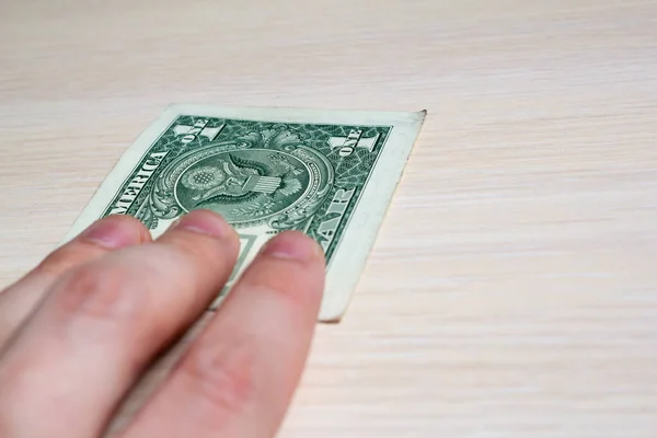 hand holds out a one-dollar bill on a light wooden table. side view. to close