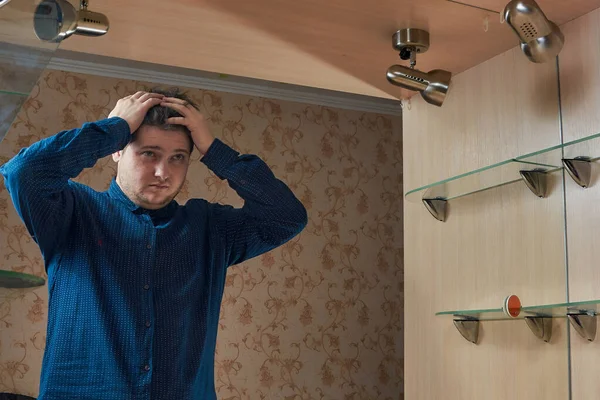 A young Caucasian man stands near a mirror holding his head looks in the mirror in a blue shirt in the home appliance