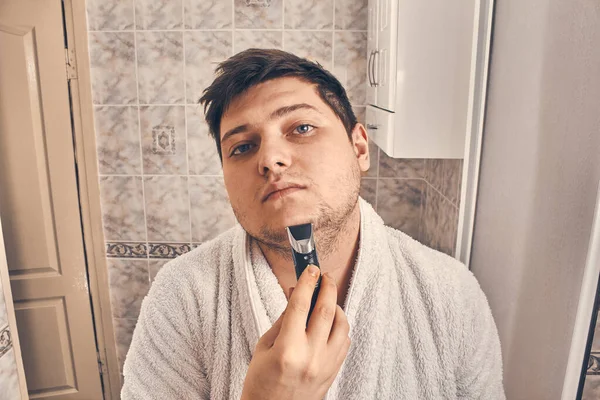 guy in a white coat shaves with a trimmer in the bathroom. close up